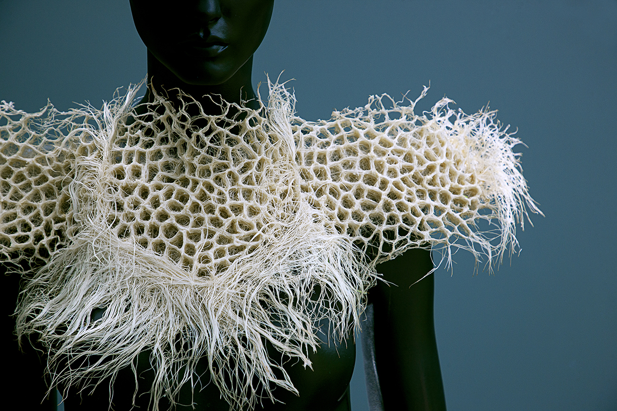 A close up of a figure wears a garment created from plant roots across its neck