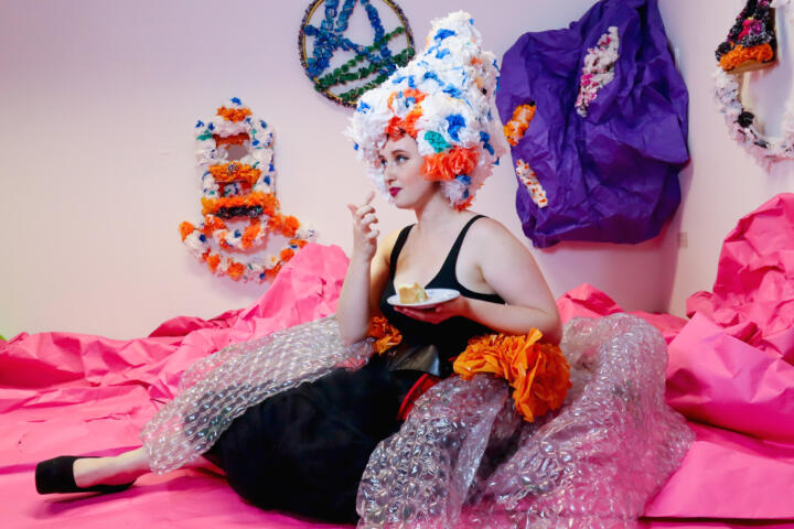 colour photo, interior. A white Drag performer sits on the floor. They are wearing a giant bubble wrap skirt with a black net underskirt and a black bralette. They have a giant wig made out of blue, white and orange plastic flowers. They are holding a piece of cake on a saucer and have a finger lifted towards their mouth.