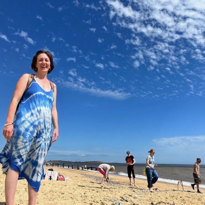 A person stands wearing a blue and white dress, smiling at the camera on a sandy beach, framed by a blue sky with some clouds. There are some people walking in the background on the shoreline.