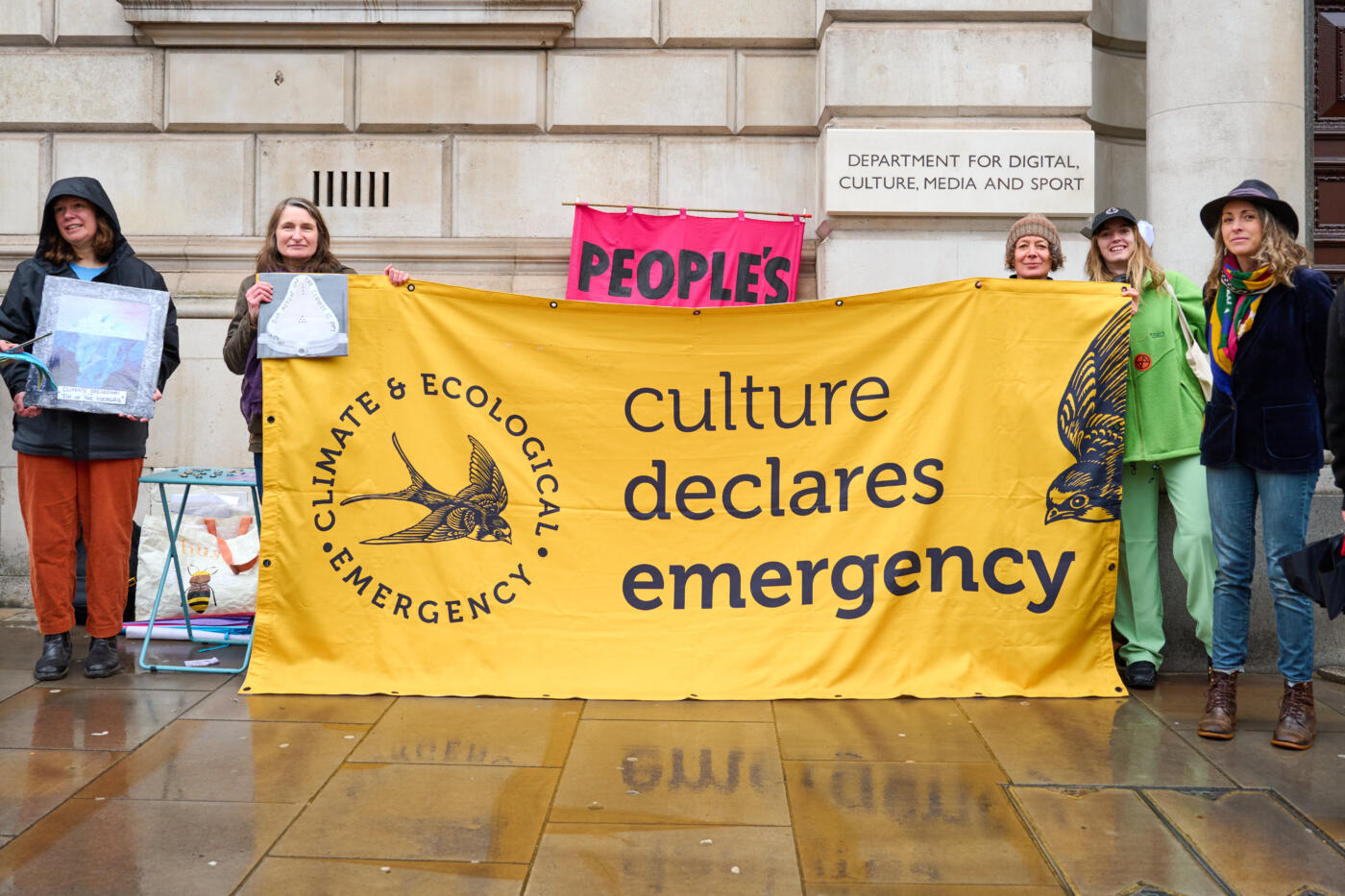 Five people stand outside of the Department for Digital, Culture, Media and Sport amongst placards, holding a large yellow Culture Declares Emergency banner.