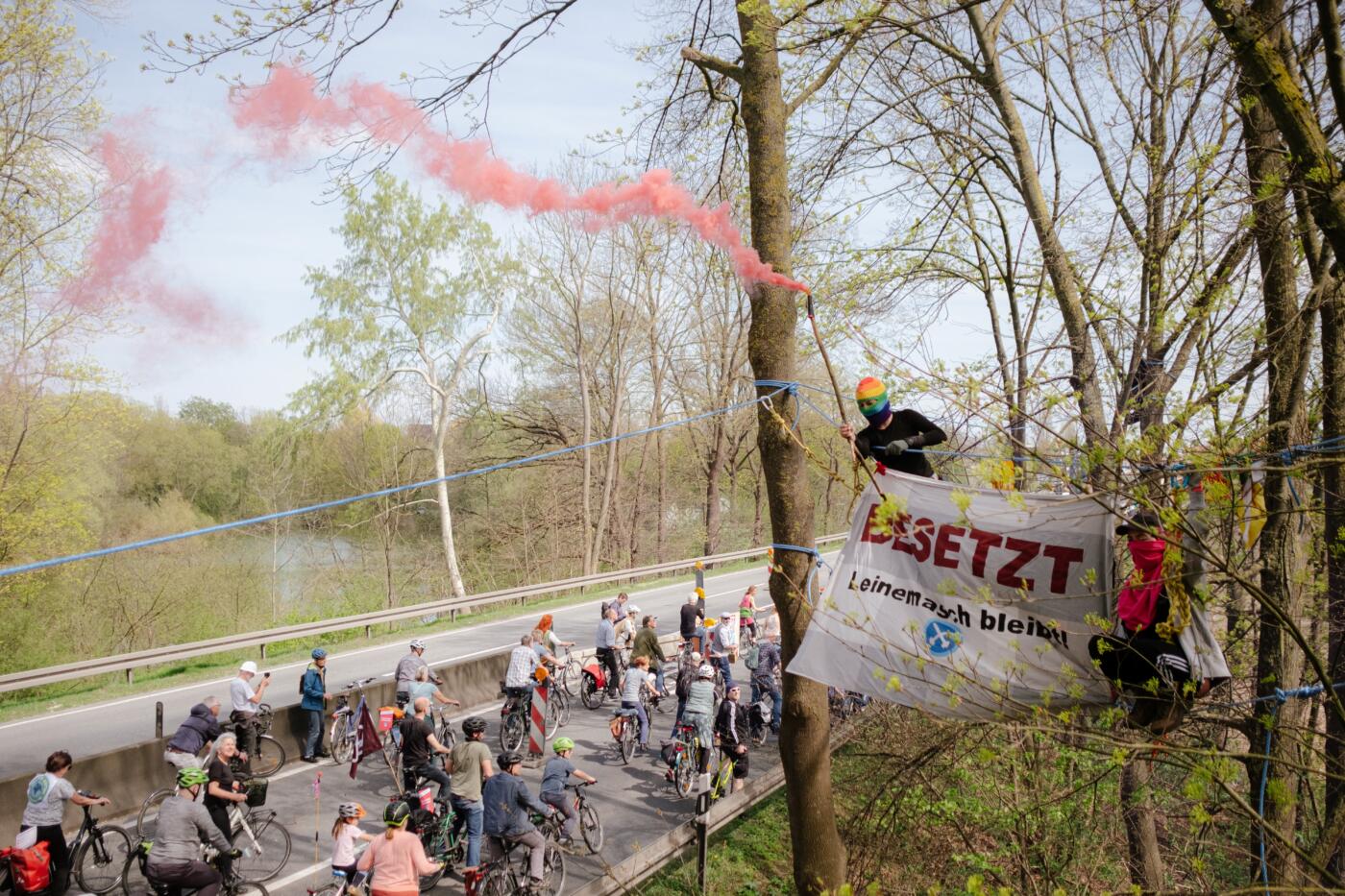 Activists on bikes cycle on the road under the canopy of trees, with activists in the trees holding up a large sign that reads "Besetzt Leinemasch bleibt"