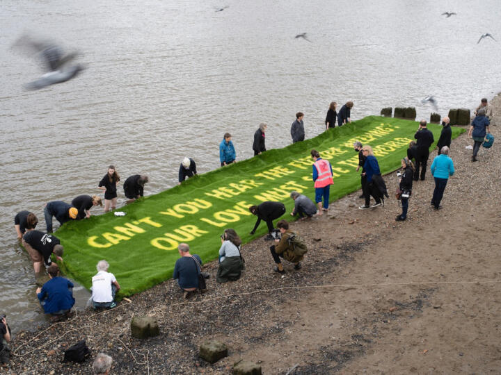 A group of people float a large grass banner on the Thames, that reads "Can't you hear the future weaping? Our love must save the world"