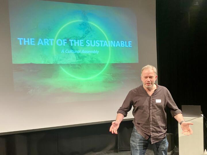 Russ Tunney stands infront of a screen that reads "The Art of The Sustainable, a cultural assembly"