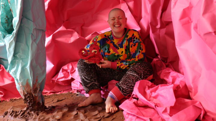 Image description: colour photo, interior. Kitt, a white, shaven headed person with a wide crinkly nosed smile, sits amongst vast sheets of pink and mint green paper crumple to form a cave -like structure around them. on the floor is wet, raw terracotta clay in rolling folds