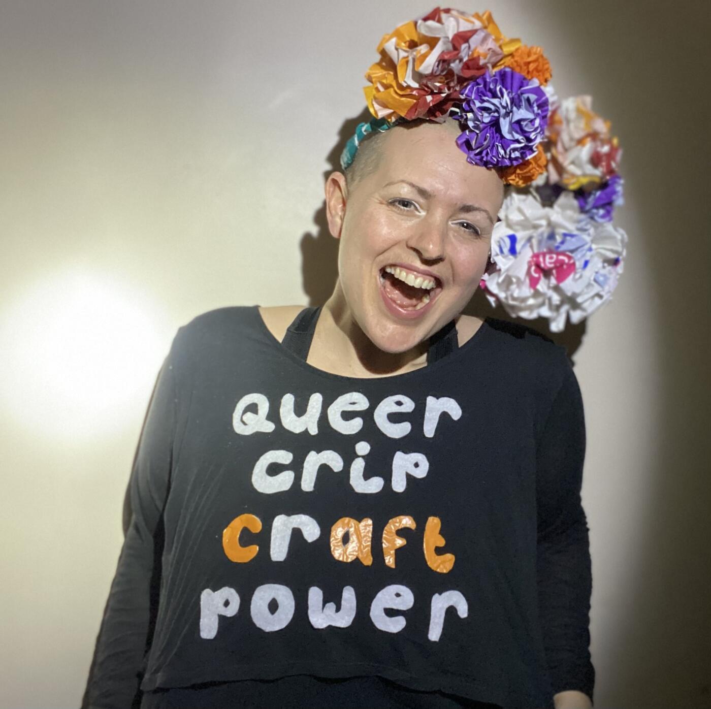 image description: Colour photo, interior. A white human with a shaved head and a big smile wears a headdress made of huge multi coloured flowers and a black long sleeved shirt on which white and orange text reads “ queer crip craft power"